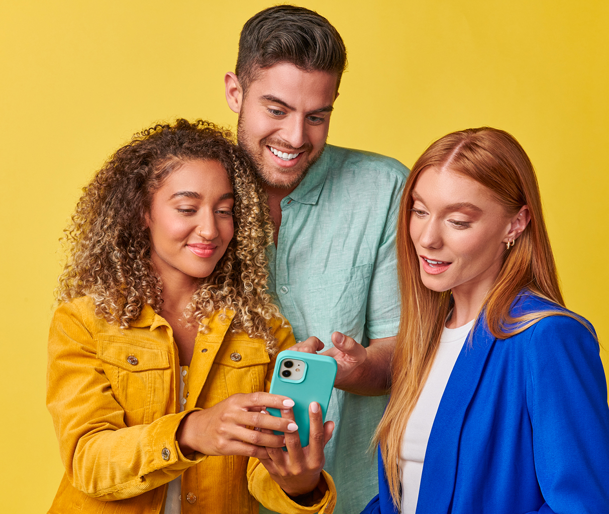 Two women and a man stand against a yellow background, smiling and looking at the smatphone. 